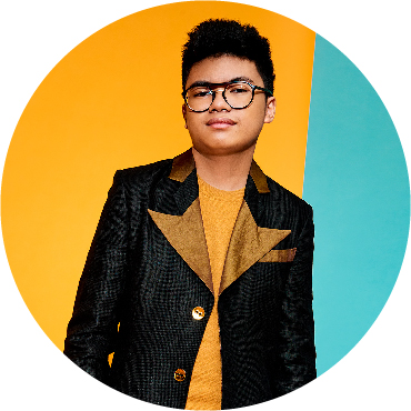 Joey Alexander stands, wearing a black jean jacket with yellow lapels, agains a half yellow, half teal background