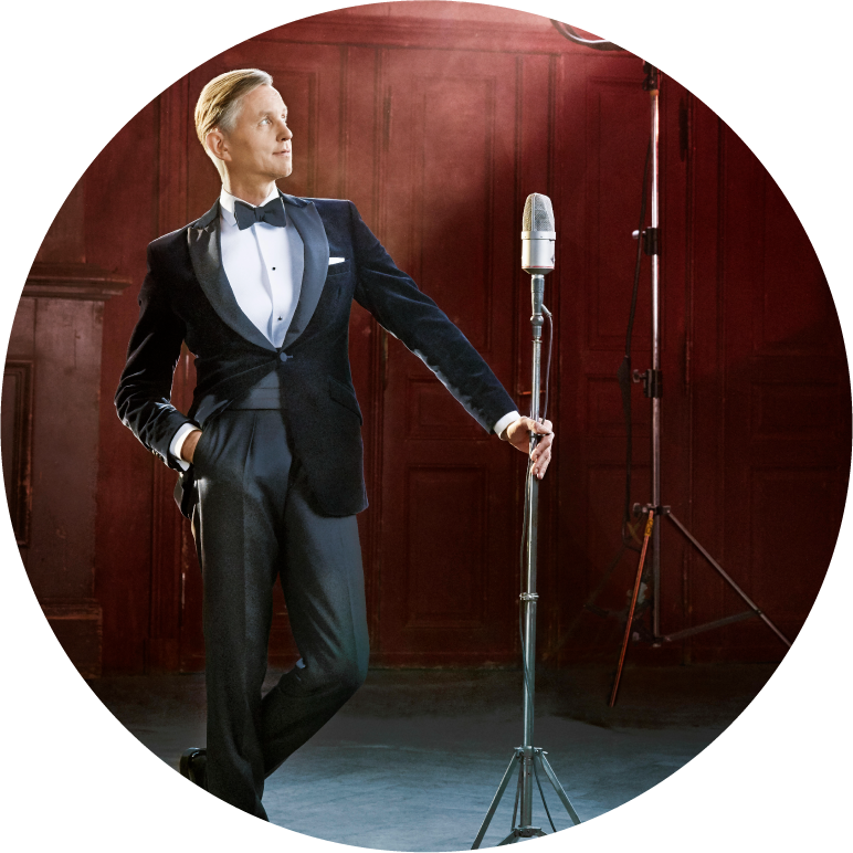 Max Raabe dressed in a tuxedo leaning against mic stand on stage in front of a red wall