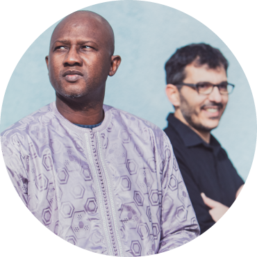 Abalye Cissoko looks off into the distance in a lavender patterned shirt who smiles and stares off. in front of Cyrille Brotto