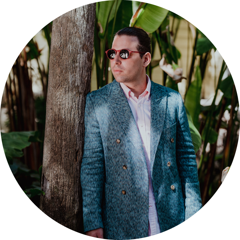 Alfredo Rodríguez wears a blue suit and sunglasses as he looks to the side and stands against a tree