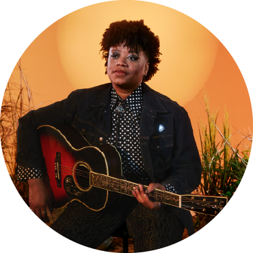 Amythyst Kiah poses with a guitar against a sunset orange background,