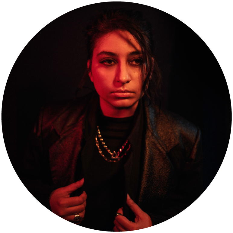 Arooj Aftab is bathed in red light, wearing a necklace over a black high neck top and a blazer over that.