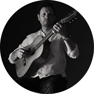 Derek Gripper holds his guitar and poses against a black backdrop