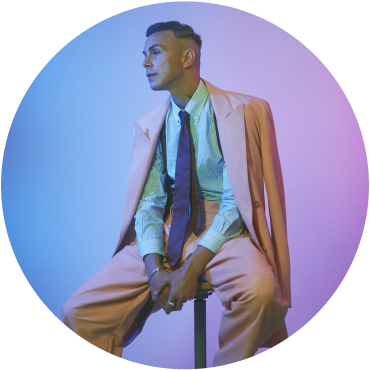 A circular image of Asaf Avidan sitting on a stool in front of a blue and purple backdrop. He has light skin and short dark hair that is shaved on the sides. He a light blue shirt with a purple tie, pink pants, and a matching pink jacket that is draped over his shoulders