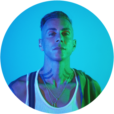 A circular bust shot of Asaf Avidan. He has light skin and short dark hair that is shaved on the sides. He looks at the camera, with half of his face in purple light and the other side is lit by green lights. He has black tattoos on his neck. He wears a white sleeveless top and black suspenders.