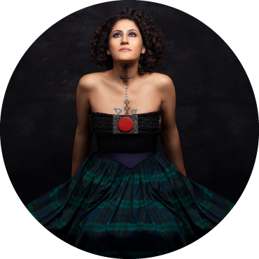 Aynur wears a black sleeveless dress with a red circle on the chest. They have black shoulder length curly hair and their face is turned upward.