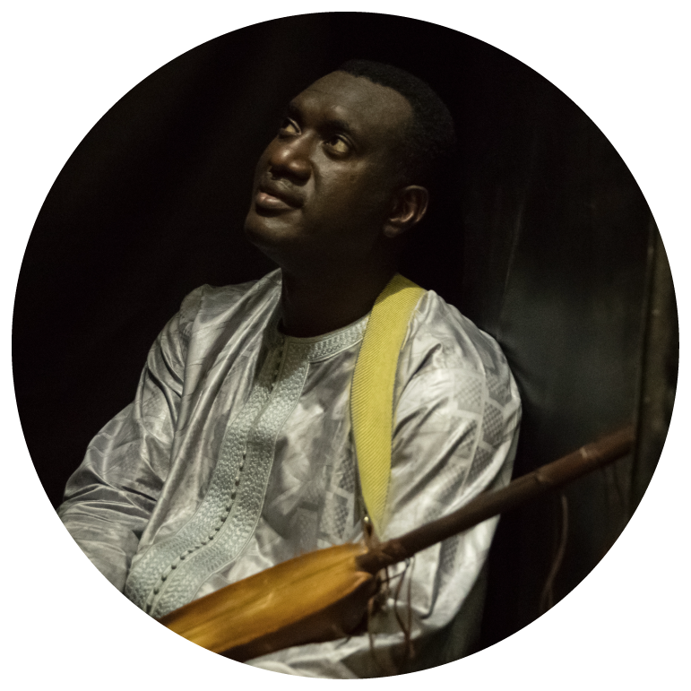 Bassekou wears a silver shirt, holds his ngoni and has his eyes closed