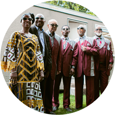 Amadou and Mariam stand next to the Blind Boys of Alabama and in front of a building. Miriam is wearing a black, white, yellow dress and a black headwrap. Amadou wears a black suit with a light blue shirt. The Blind Boys wear red suits. They are dark-skin and wear sunglasses.