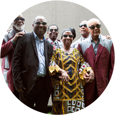 The Blind Boys of Alabama wear red and gray suits. Miriam and Amadou stand in front of them. Amadou wears a black suit and a light blue shirt. Miriam wears a yellow, black, and white dress and a black headwrap. They have dark skin and wear sunglasses.