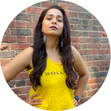 Purnima stands with her arms on her hips with her long brown hair in waves down her yellow tank top that has the words BollyX on it