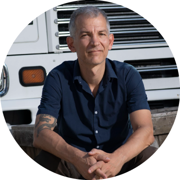 Brad Mehldau wears a blue shirt and brown pants and faces forward against a white vehicle in the background