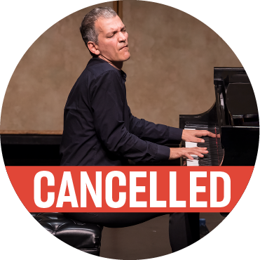Brad Mehldau in black attire playing grand piano passionately on stage with a poppy colored "cancelled" banner over the photo