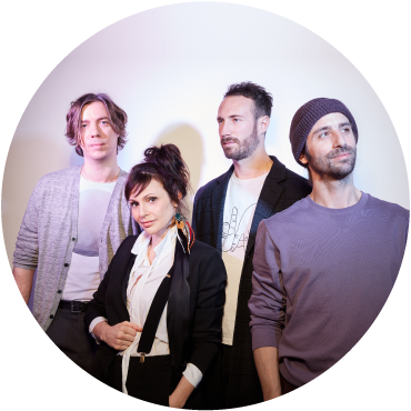 A circular image of the 4 members of Caravan Palace in front of a light background. The 1st person on the left has light skin & wavy blonde jaw-length hair. He has a white shirt & a light blue sweater. The 2nd person is shorter with dark hair in an updo. She has a white button-up shirt & a black jacket. The 3rd person is taller, with light skin & short dark hair with a short beard. He wears a black jacket over a white t-shirt. The last person has light skin with beard a blue shirt.