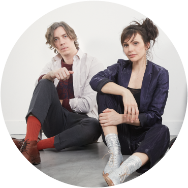 2 members of Caravan Palace sit on the floor in front of a white background. The 1st is light-skinned with blonde hair that comes to his jaw. He wears an open white shirt over a dark red t-shirt, black pants, red socks, and brown shoes. Both knees are bent with one on the floor. He leans his hand on the other knee. The 2nd person has light skin and dark hair that is in an updo. She wears a black jacket, shirt, and pants and silver pants. She leans her elbow on one bent knee.