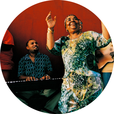 Cesaria Evora is smiling with their mouth open and their hands are raised. They are wearing a green and yellow patterned dress and are standing in front of a band.