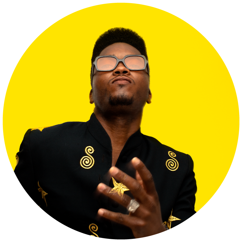 Cimafunk stands against a yellow background. He wears a black jacket with gold detailing on the lapel. He wears black sunglasses and his left hand is raised over his chest.