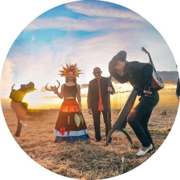 The sun sets in the background of a field of straw, with musicians in black hats dancing around a female holding an animal horn