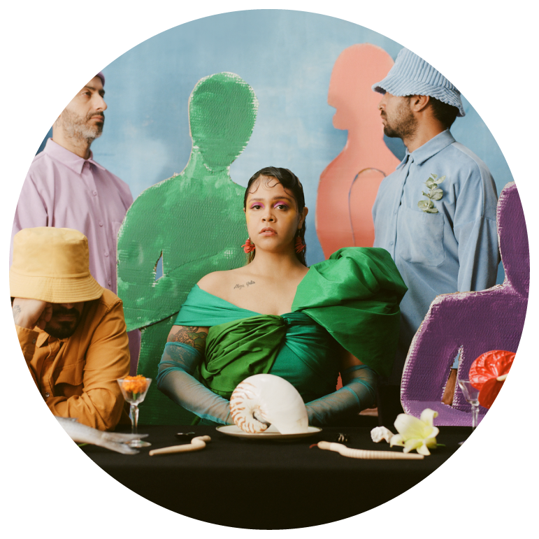 A person with straight dark brown hair sits at a table wearing a green dress. They are surrounded by 3 others, to her right a person sits with a yellow outfit and hat with another person in pink standing behind them and a person in a blue hat and shirt stands to theirleft