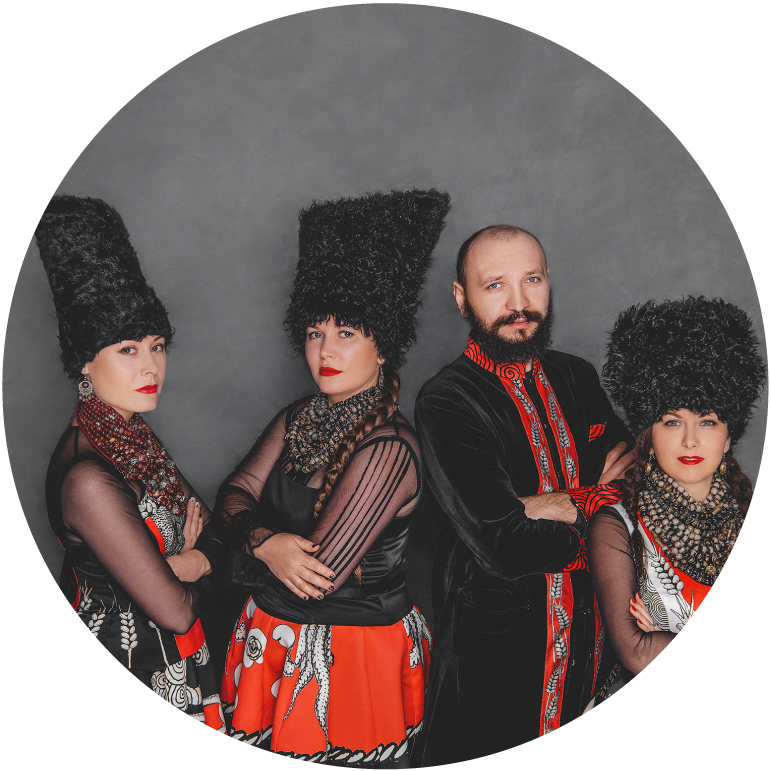 Four members of DakhaBrakha stand in front of a gray background. They are wearing traditional black and red clothes.