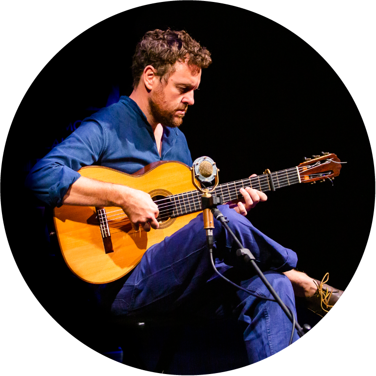 Derek Gripper wearing blue pants and a blue button up top, playing the guitar with his right leg crossed over the left