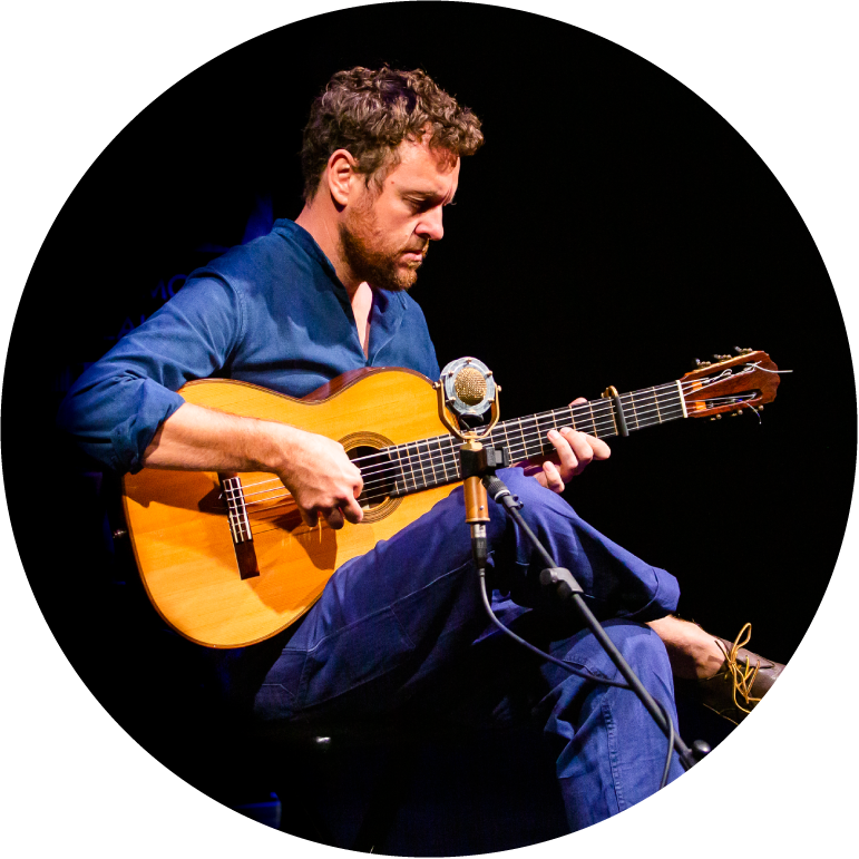 Derek Gripper sits in front of a black background. He is light-skinned with short brown hair and beard. He wears blue shirt and pants and plays an acoustic guitar that is propped up on his leg.