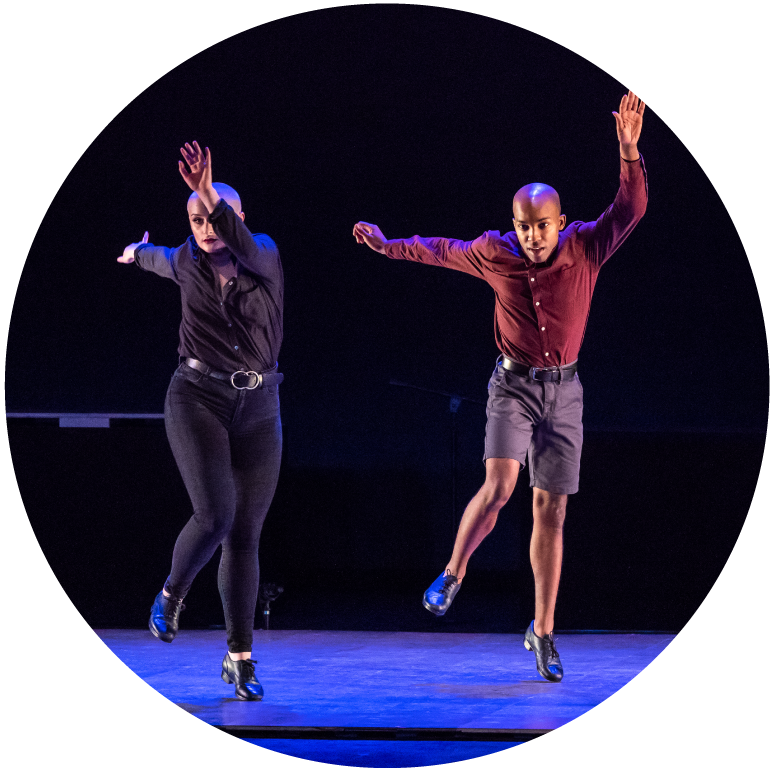 Two dancers wearing button up shirts have left arms extended in front and right arms extended behind them. They are mid-jump.