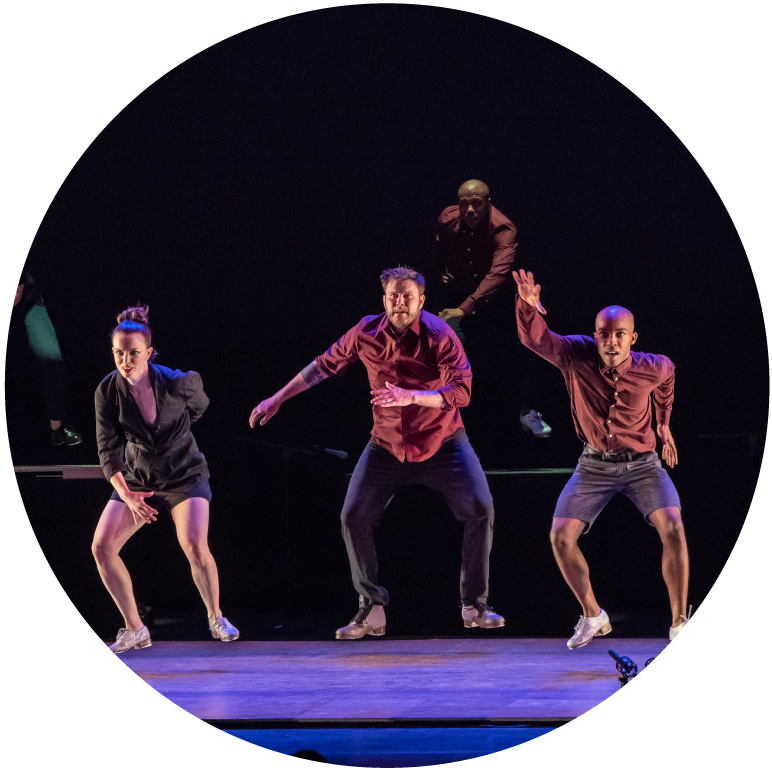 Three dancers face forward with knees bent and arms at the ready. The background is dark.