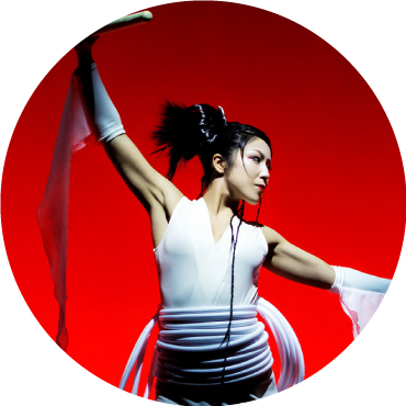 Drummer poses in white clothing with hands stretched to the side against a red backdrop