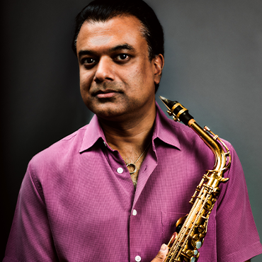 Rudresh Mahanthappa in pink shirt holding saxophone while giving the camera a cool gaze in front of a grey background