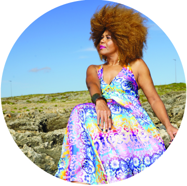Fantcha sitting on the beach with her legs crossed and her right arm crossed over the leg, her hair is big, curly, beautiful and blowing in the wind