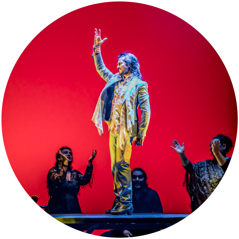 Farruquito stands on a platform surrounded by other performers. He wears a white suit and faces to the right with his right hand raised above his head 