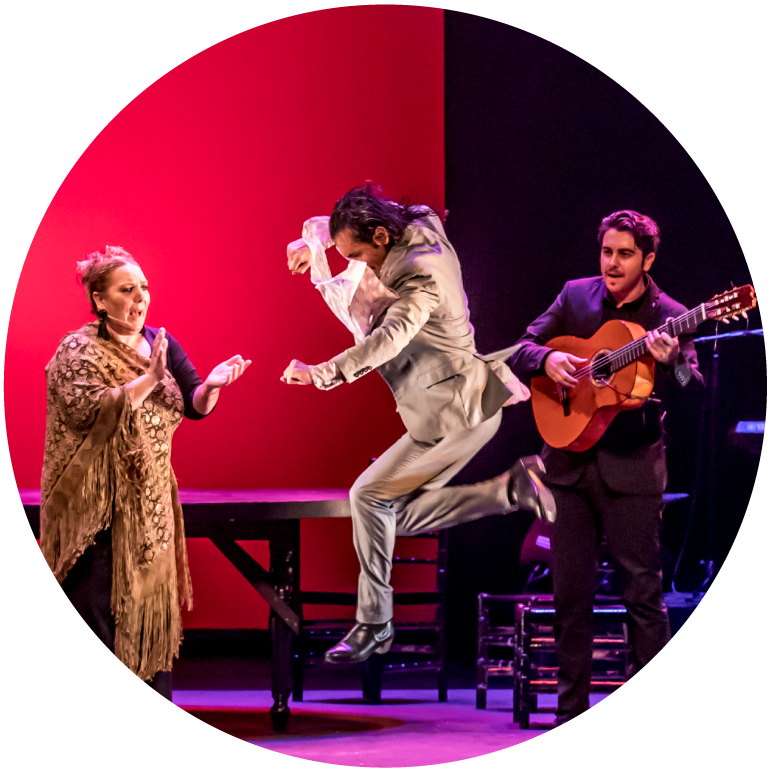 Farruquito is mid jump wearing a white suit. A female dancer in a dress is on his left and a male guitarist is on his right