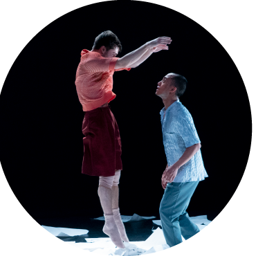 Two dancers with short black hair. One is wearing a blue shirt and pants and is looking up at the other, who is wearing an orange shirt, red pants, and is jumping with their arms raised. They are in the middle of a pile of papers with various words on them.