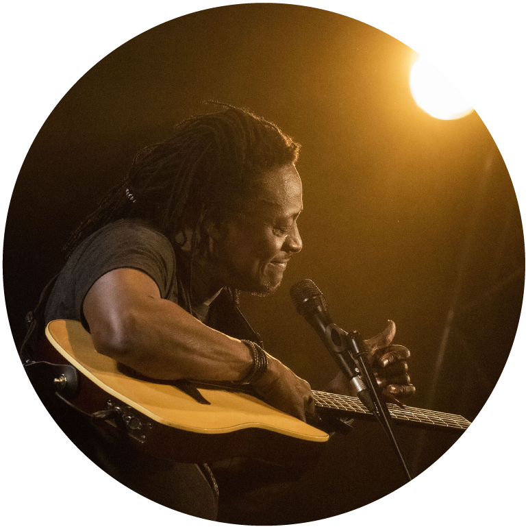 Habib holds his guitar up almost parallel to the ground with his hand on its neck. A mic is in front of him and he is bathed in a warm yellow from a spotlight in the top right of the photo