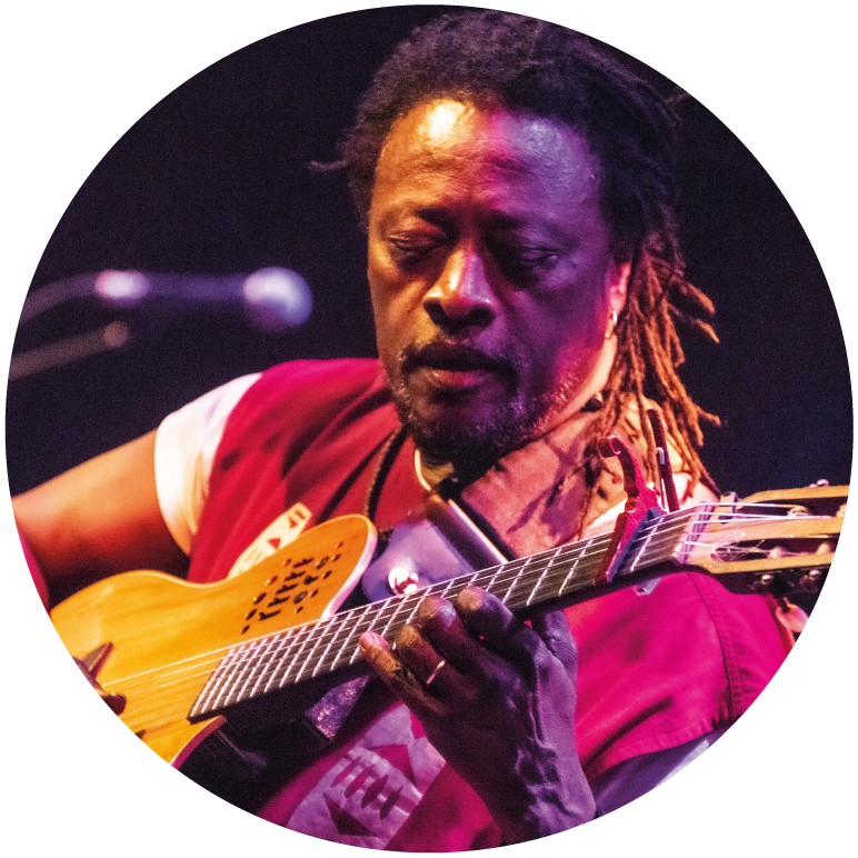 Habib holds his brown guitar and looks down on it. He wears a pink top, hair in dreadlocks, a mic in front of him
