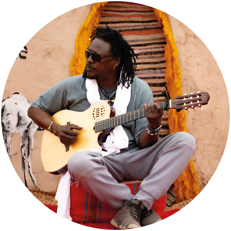 Habib sits on steps in front of a door to a clay hut. He holds his guitar and wears grey pants, a mint green top with the sleeves rolled up and a white scarf and black sunglasses