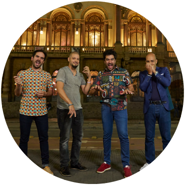 Harold López-Nussa's quartet poses on a street in front of Theatro Municipal in São Paulo, Brazil at night. The quartet is wearing jeans and pretending to play instruments.