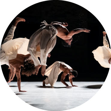 Four dancers are all at different points of being mid-flip. They are all wearing flowy white pants and no shirt.