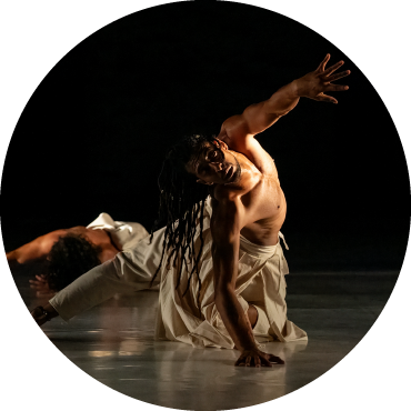 A person with long black hair is wearing flowy white pants and no shirt. They are contorted and twisted to face the camera, reaching out in front of them.