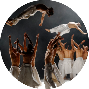 Two dancers are being tossed in the air by separate groups of dancers. Everyone is wearing flowy white pants and no shirt.
