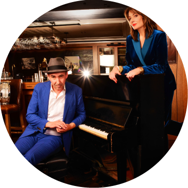 The person on the left has short blond hair and is wearing red pants and a green floral blouse. They are singing into a microphone. The person on the right is wearing a black suit and hat and is dancing while facing away from the camera.