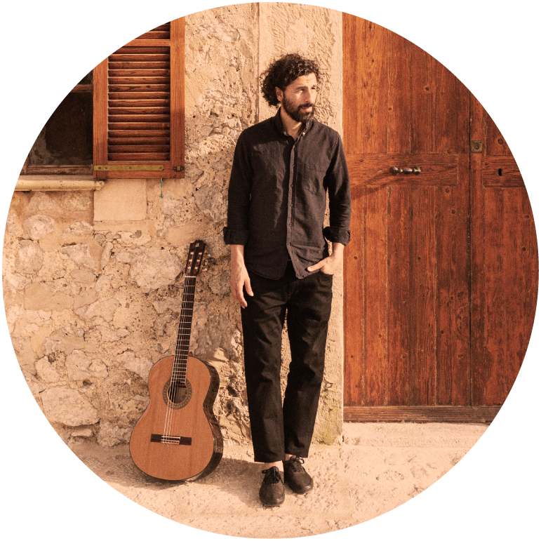 José González stands against a brown backdrop with his guitar, as he looks off to the side