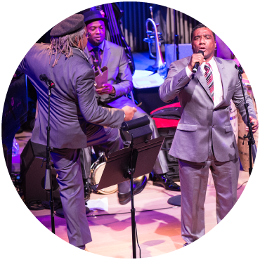 Juan de Marcos dressed in a grey suit turns his back while two performers in grey suits sing and play an instrument.