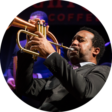 A member of the Afro-Cuban All Stars is playing a trumpet. They are wearing a black suit and have short black curly hair.