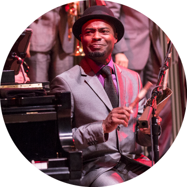 A member of the Afro-Cuban All Stars is playing a percussion instrument and sitting next to a piano. They have a short black beard and are wearing a silver suit with a black fedora on their head.