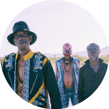 Jupiter Bokondji Illola in the foreground wears a black hat and patterned top with his bandmates of Okwess in the background against the backdrop of a mountain.