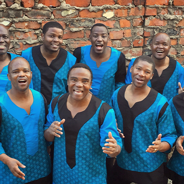Members of Ladysmith Black Mambazo stand in front of a brick wall wearing matching blue and black shirts with smiles on their faces