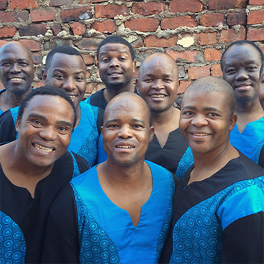 Members of Ladysmith Black Mambazo stand in front of a brick wall wearing matching blue and black shirts with smiles on their faces