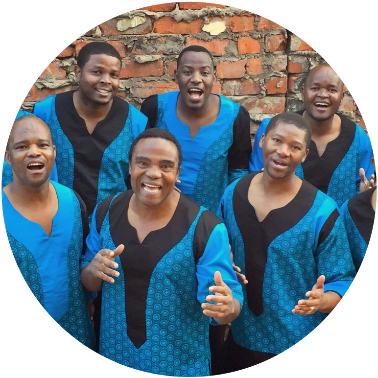 6 members look at the camera and stand in front of a red brick wall. They wear blue and black robes. 