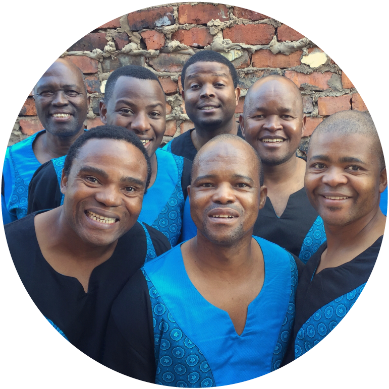 The seven members look at the camera and stand in front of a red brick wall. They wear blue and black robes.